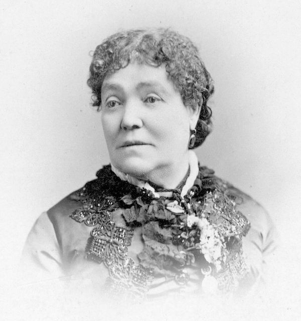 Eilley Bowers in 1863, at the height of her fortune: a studio portrait of a woman with curled hair, in a dark dress trimmed with lace and beadwork. Black-and-white photograph.